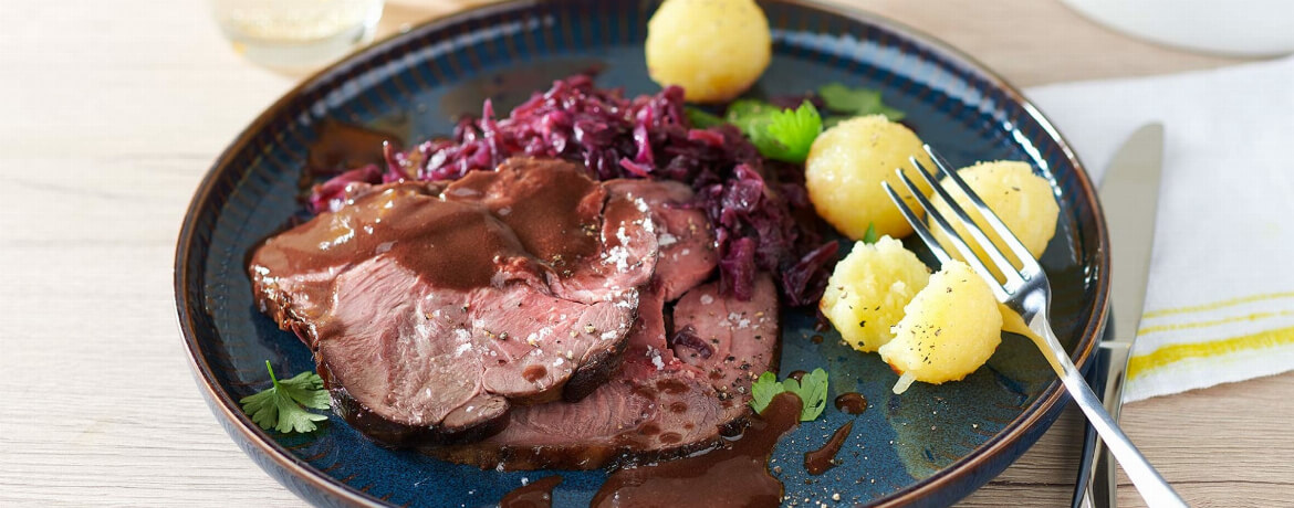 Rehrollbraten mit Rotkohl auf einem Teller mit Petersilie sowie einer Apfel-Rotkohl-Mischung und Kartoffelknödeln